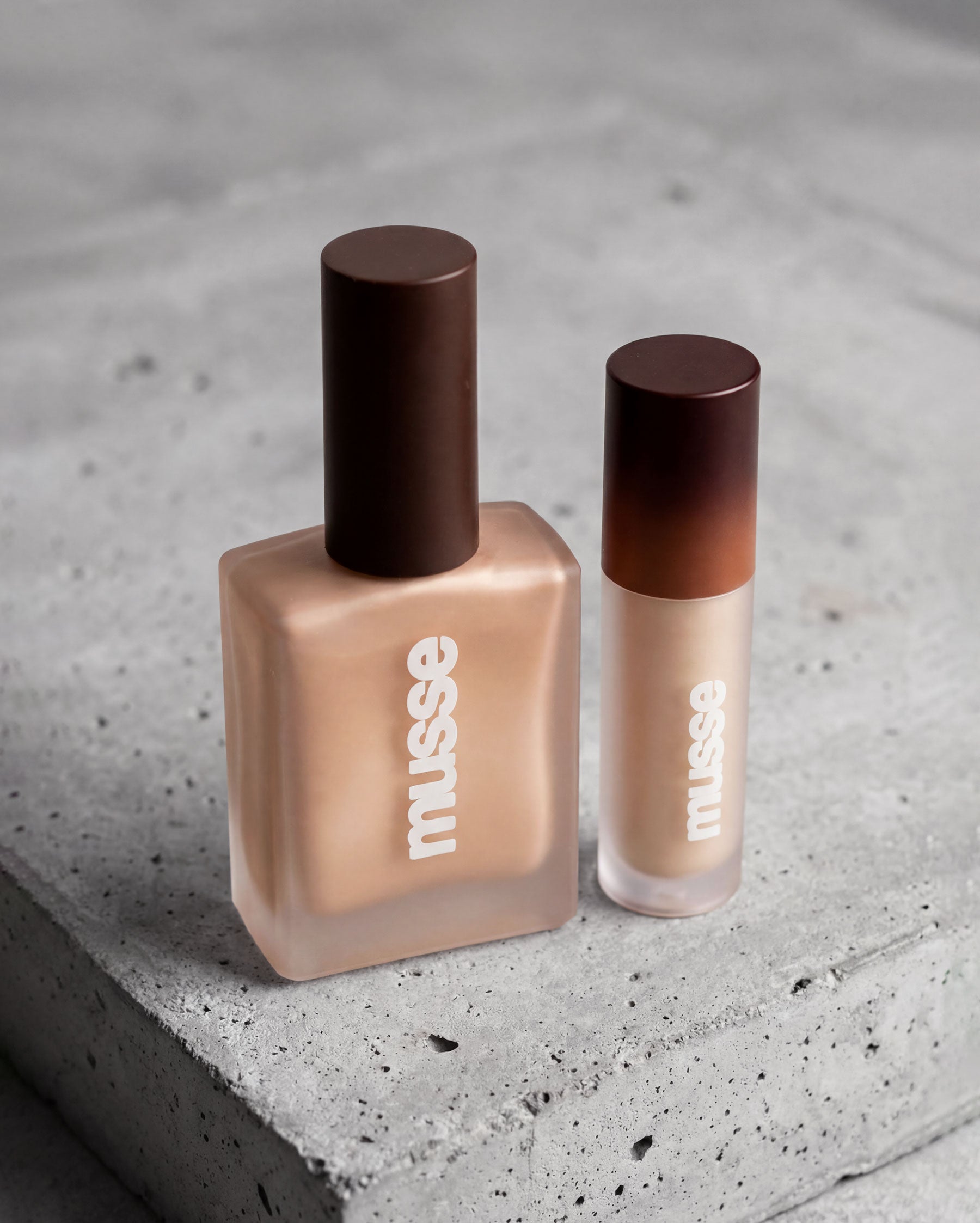 Pack Glow Musse Cosmetics con iluminador corporal líquido y mini iluminador, tonos dorados sobre fondo de concreto.