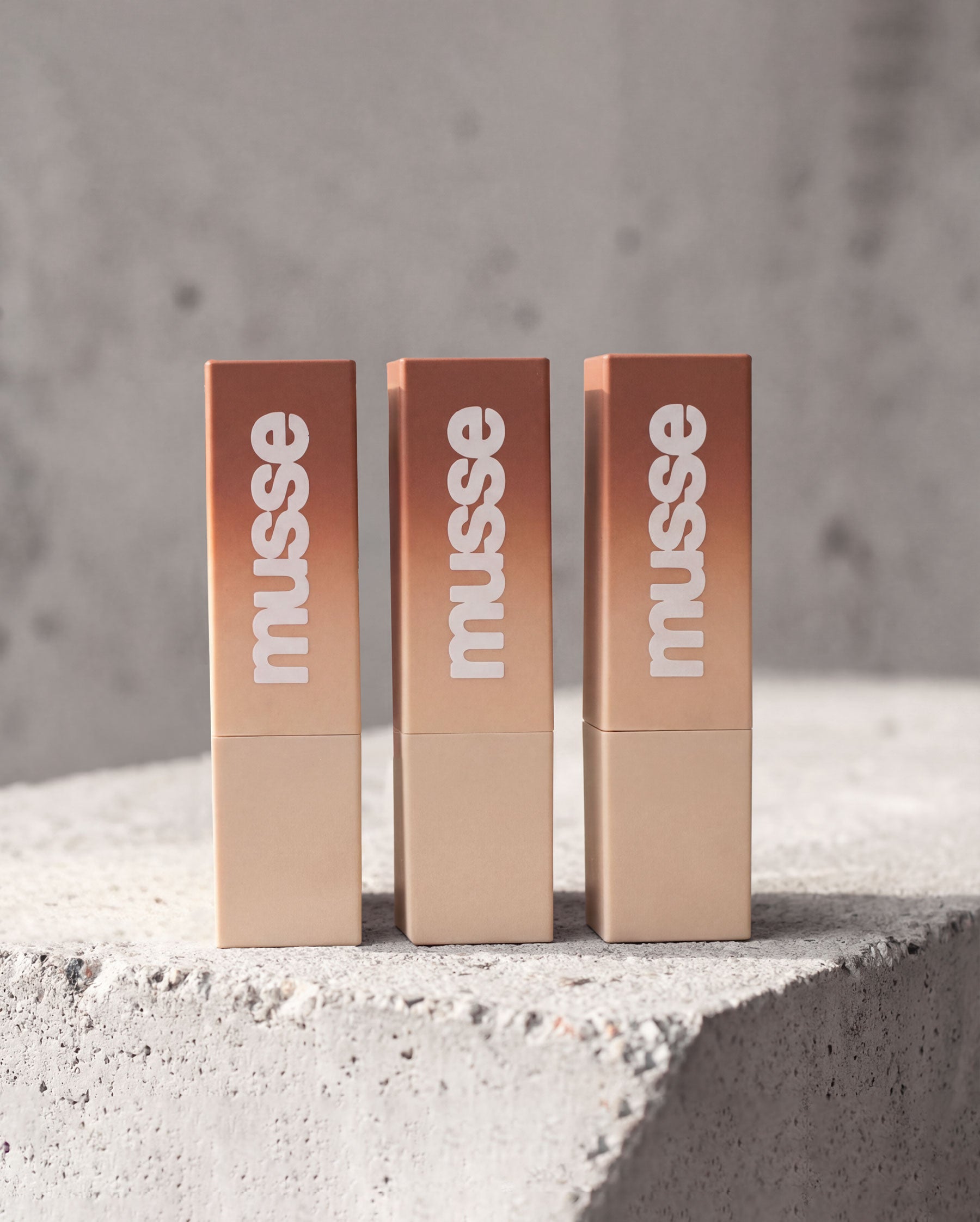Tres labiales Musse en tonos cálidos sobre una superficie de concreto, con envase beige degradado y logo blanco