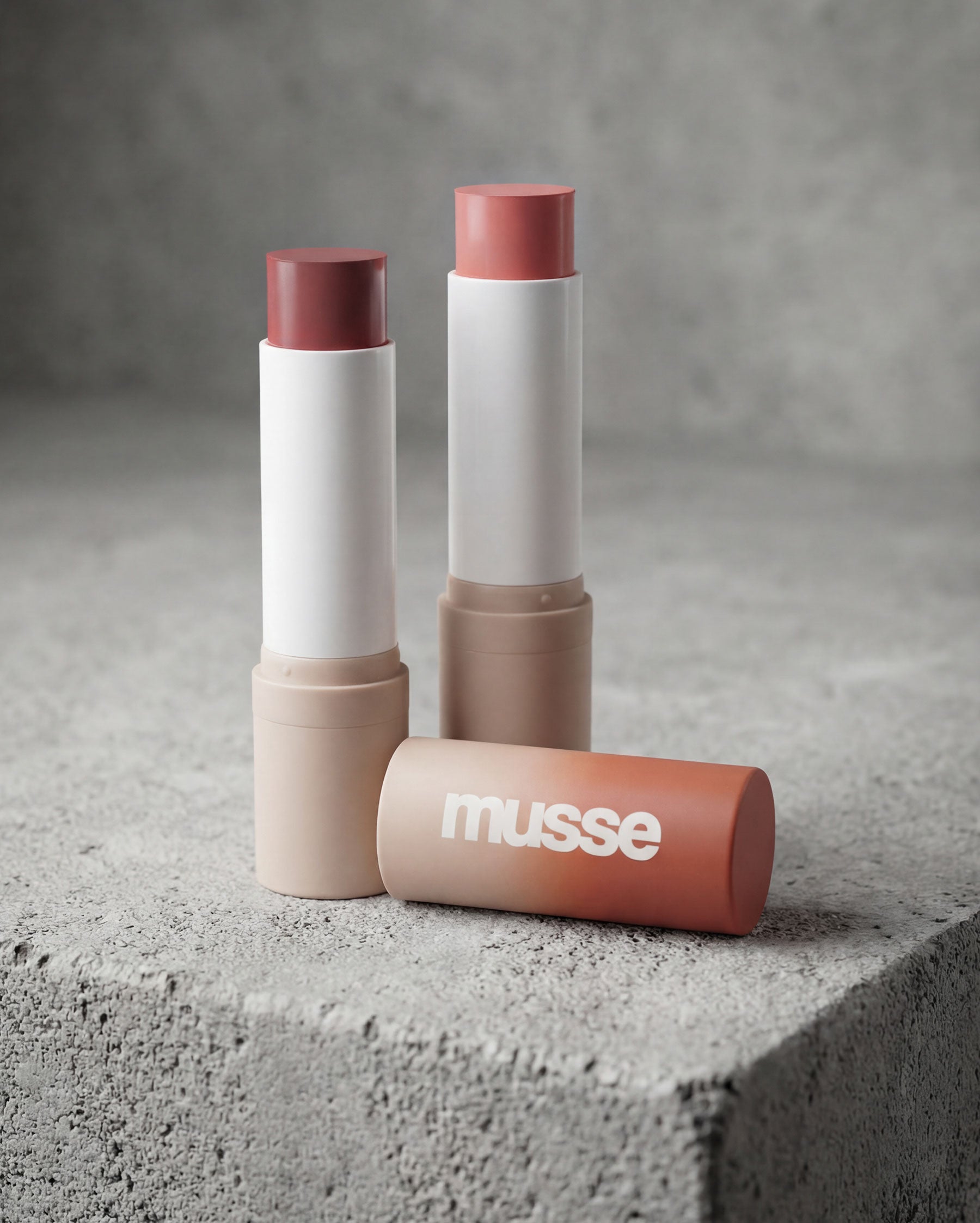 Rubores en barra Musse Cosmetics en tonos cálidos, fotografía de producto sobre superficie de cemento.